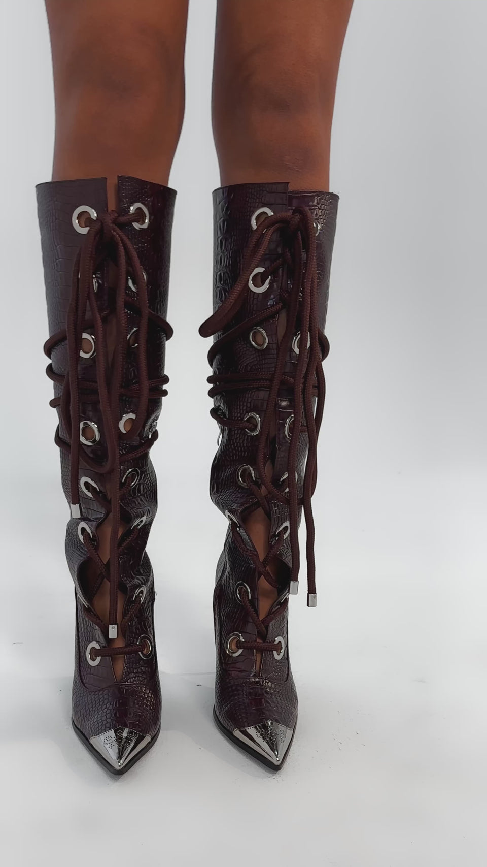 Video of woman modeling the AKIRA GOLD PANTOUM CHOCOLATE CROC STILETTO BOOT, featuring dark brown faux-crocodile leather, lace-up front with large silver eyelets, pointed metal-capped toes, and high stiletto heels.
