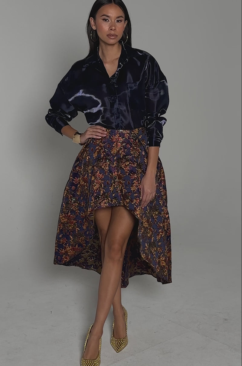 Video of woman modeling the STAR OF THE NIGHT BROCADE MIDI SKIRT, a dark floral high-low skirt with a satin finish and metallic detailing, paired with a navy blouse, gold bracelet, and gold textured high heels, standing against a neutral background.