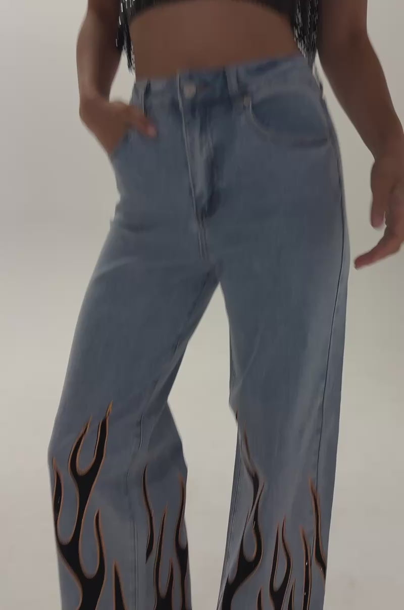 Video of a woman modeling the ON FIRE RELAXED DENIM—light blue high-waisted, wide-leg jeans featuring bold black flame designs and rhinestone fire detailing on the legs.