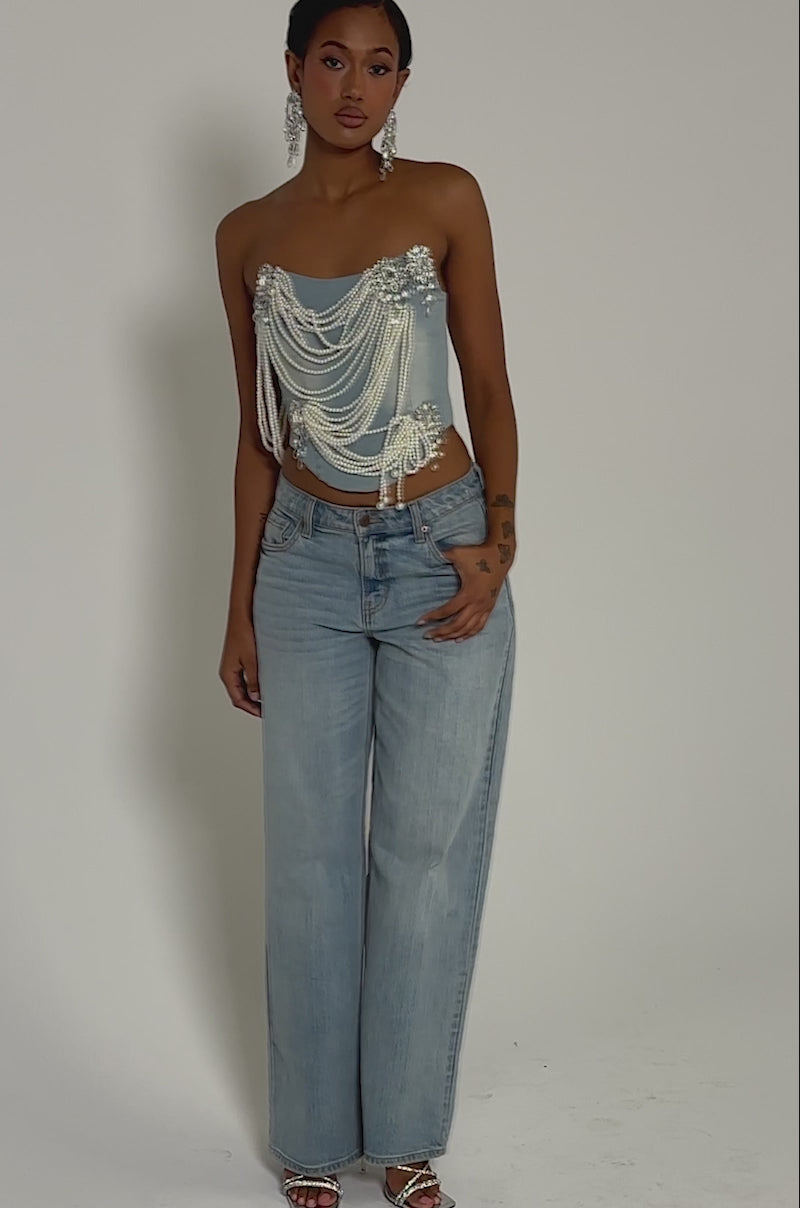 Video of woman modeling the PEARL QUEEN DENIM CORSET adorned with pearl chains and silver accents, paired with light blue jeans, large dangling earrings, and a tattoo on her left forearm.
