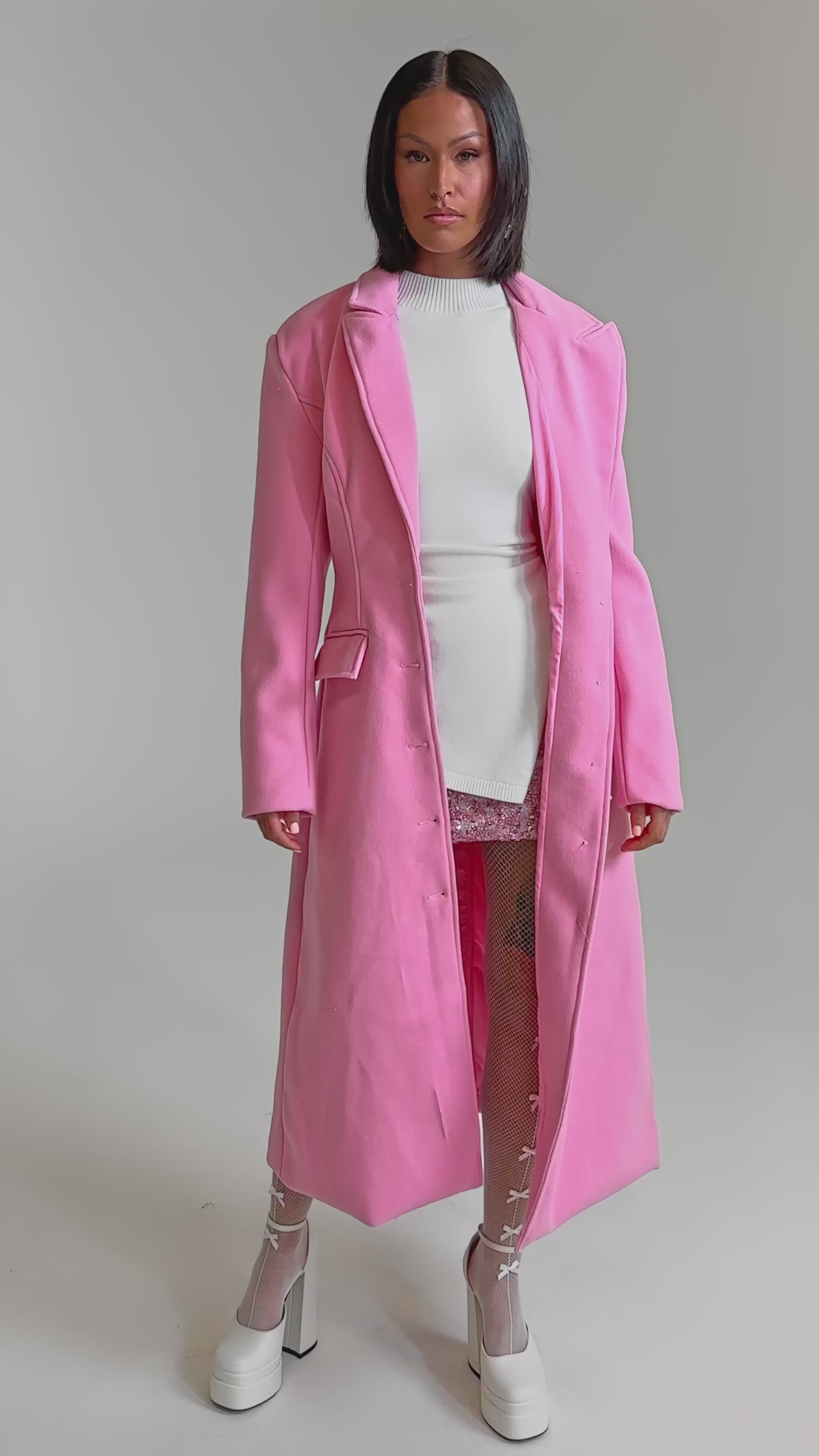 A video of a woman with a sleek bob in the SWEET THING WOVEN TRENCH in pink over a white top, pink sequin mini skirt, bow-accented fishnet tights, and white platform heels, standing confidently against a plain background.