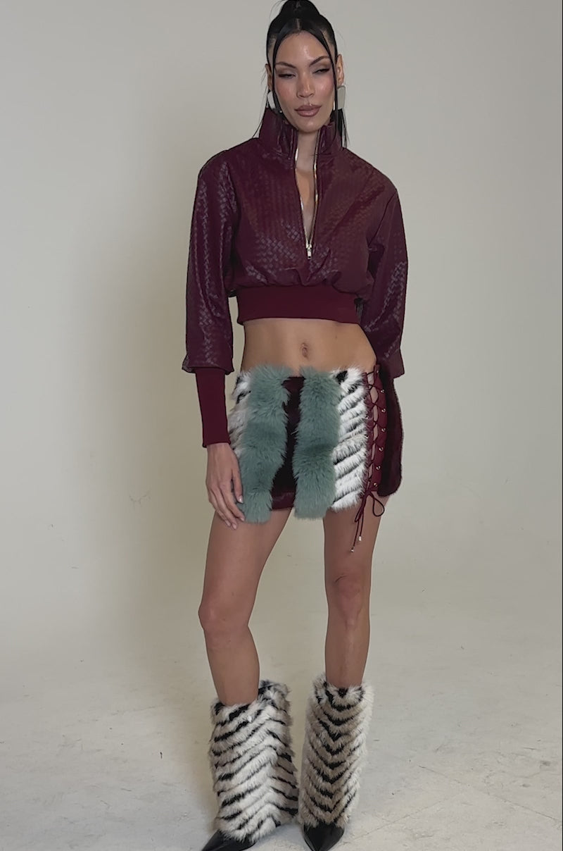 A video of a model wearing a burgundy textured crop jacket, the HAVE IT ALL PANELED FUR SKIRT with teal and fur stripes, and matching furry leg warmers featuring black stripe patterns.