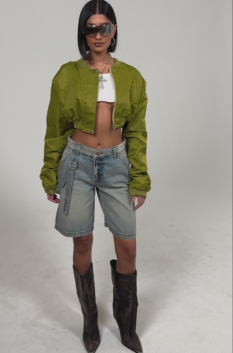 Video of woman modeling the BABY ITS YOU DENIM SHORT, a shiny olive-green cropped jacket, white crop top, tall pointed brown boots, and sunglasses.