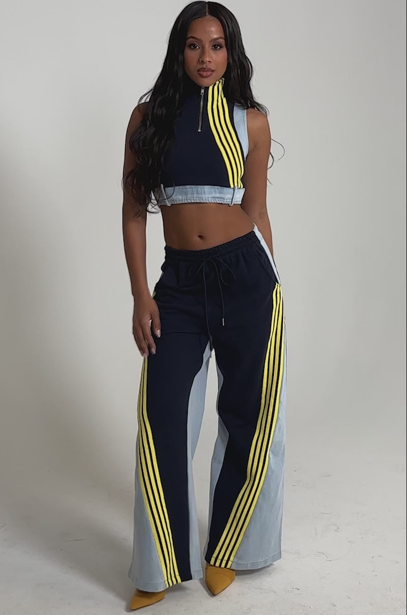 Video of woman modeling the HEADS UP DENIM WIDE LEG JOGGER IN NAVY, styled with a sleeveless navy and light denim crop top featuring a yellow stripe. The joggers have an elastic waistband and are paired with yellow pointed-toe heels.