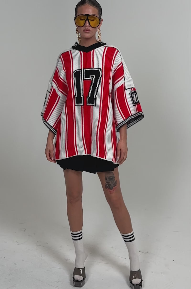 Video of woman modeling the THROW SUM MO KNIT JERSEY TOP, a red and white striped varsity sweater with "17," paired with black shorts, tall white socks with black stripes, silver platform heels, yellow sunglasses, and bold earrings.