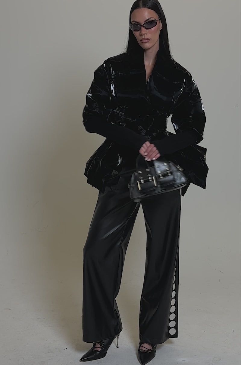A video of a woman wearing black sunglasses, pointed heels, wide-leg black leather pants, holding a small handbag—and the RIB SLEEVE PEPLUM PUFFER IN LIQUID VELVET.