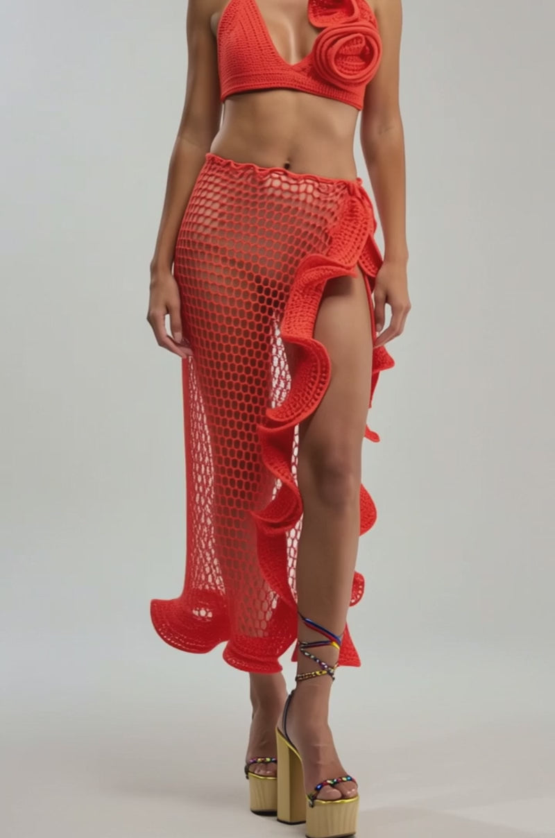 A video of a woman in the BEACH BABE CROCHET BRALETTE in bold orange with a large ruffle, paired with a matching asymmetrical skirt, platform sandals, and green statement earrings against a plain backdrop.