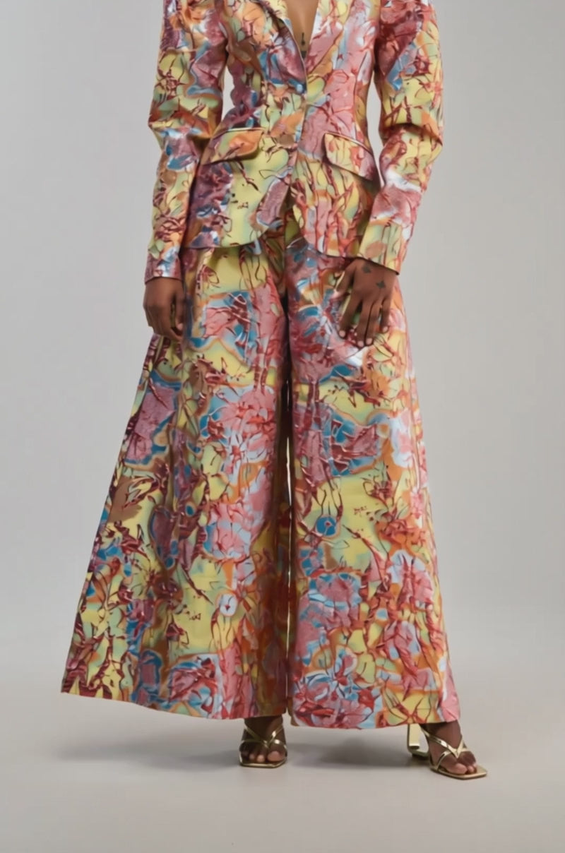 Video of a woman modeling the LUCY BROCADE BLAZER IN BLUE MULTI, a vibrant suit featuring structured shoulders and wide-leg pants with a striking marble pattern in yellow, red, and blue tones. 