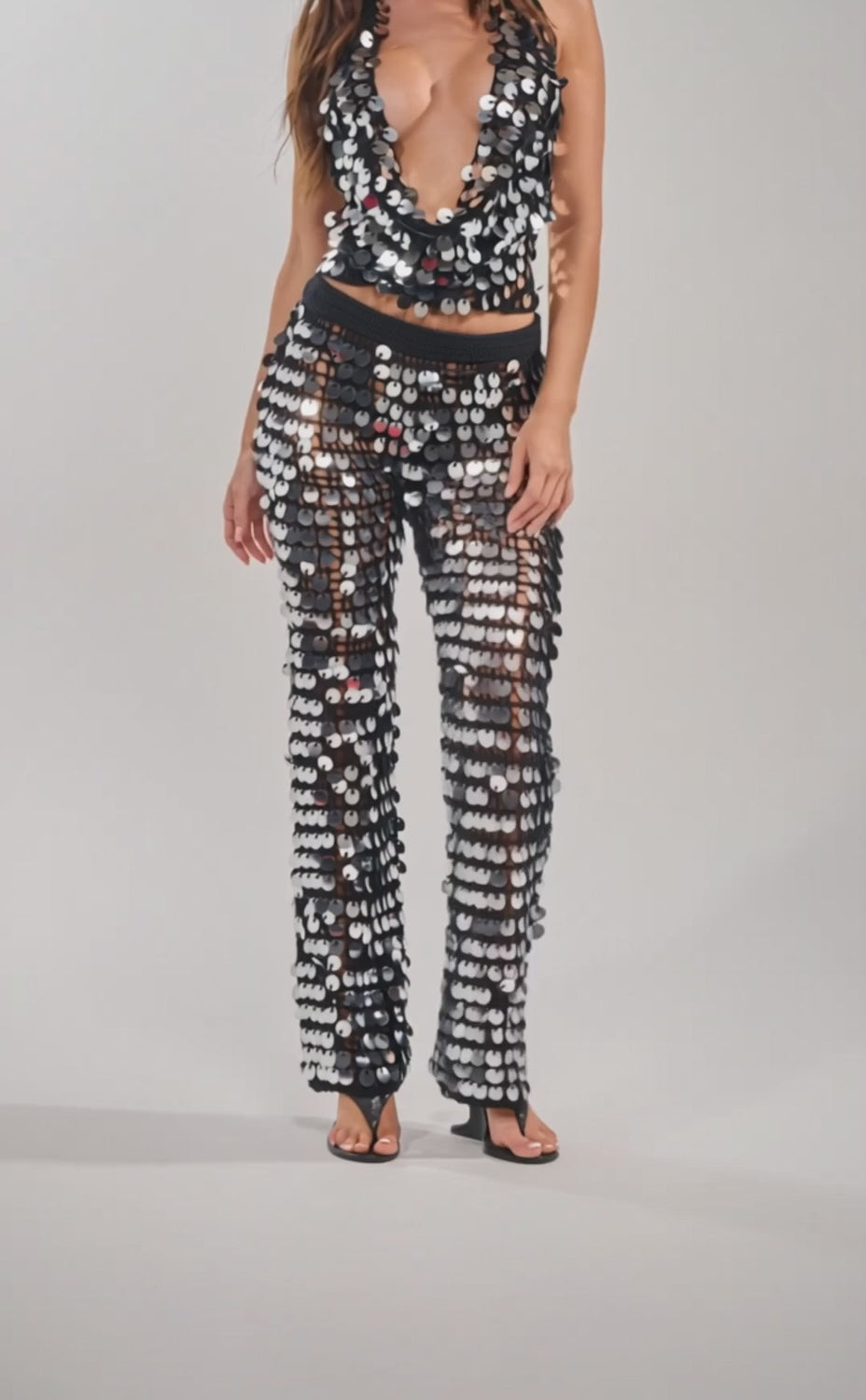 A video of a woman wearing the GIVE IT TO ME BABY CROCHET PANT and a matching halter top, both draped and low-cut with shiny silver sequin accents. 