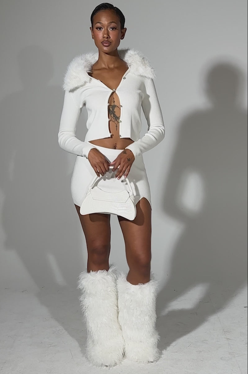 A video of a woman modeling the READY FOR THE SNOW CARDIGAN WITH FUR TRIM, paired with a matching mini skirt and tall white furry boots. She carries a textured white handbag and stands against a plain light background.