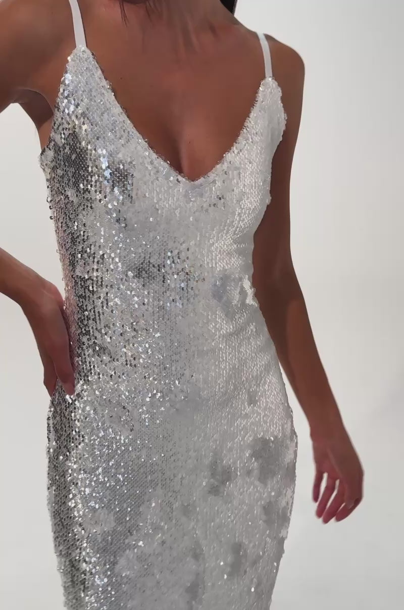A video of a woman wearing the WORTH THE STARES EMBELLISHED MAXI DRESS—a shimmering white sequin mermaid gown with thin straps and a flared faux fur hem.