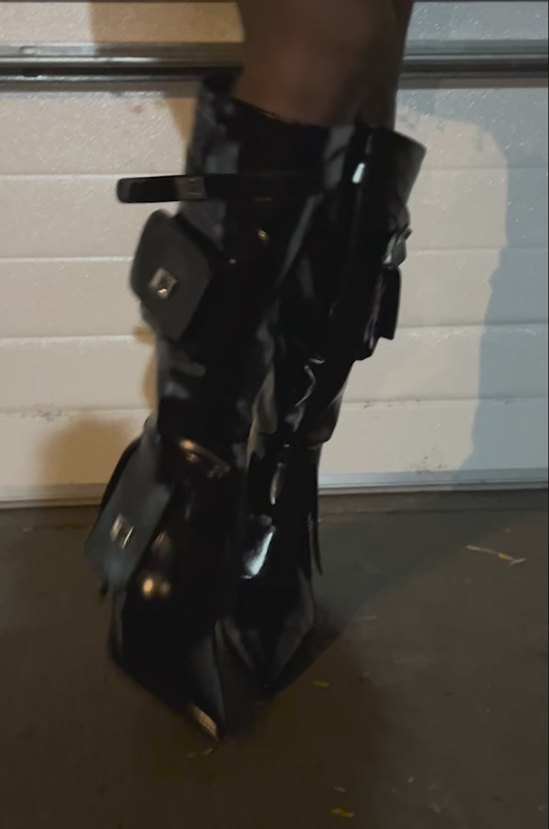 Video of woman modeling the AZALEA WANG WATERS BLACK STILETTO BOOT WITH POCKETS—shiny, faux leather knee-high boots with pointed toes, purse pockets, and silver zippers—paired with a skirt detailed in metal rings.
