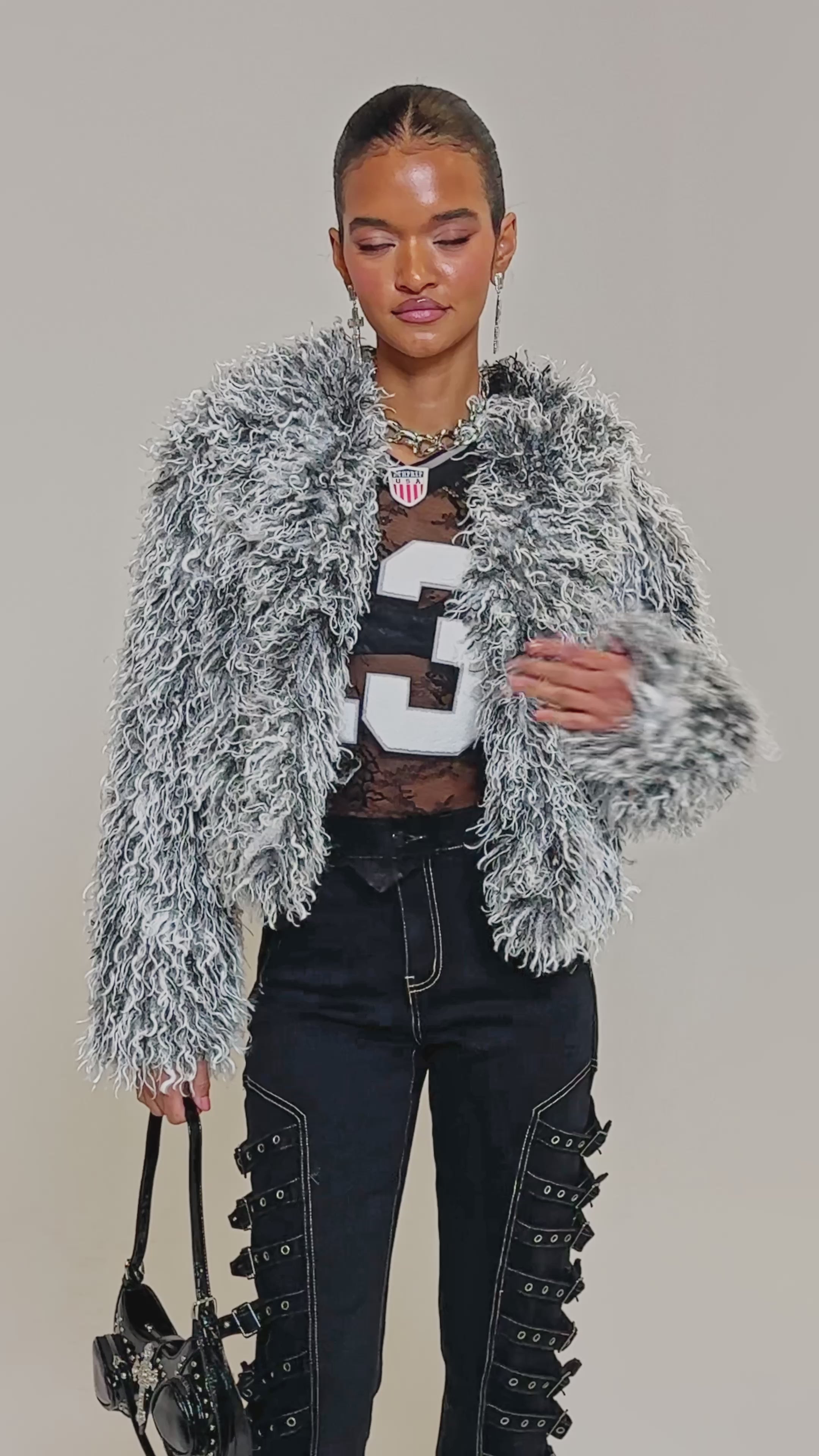 Video of woman modeling the RENDEZVOUS SHAGGY FAUX FUR cropped coat in gray and black, paired with black pants featuring visible stitching and buckles, plus large cross earrings, standing before a plain background.