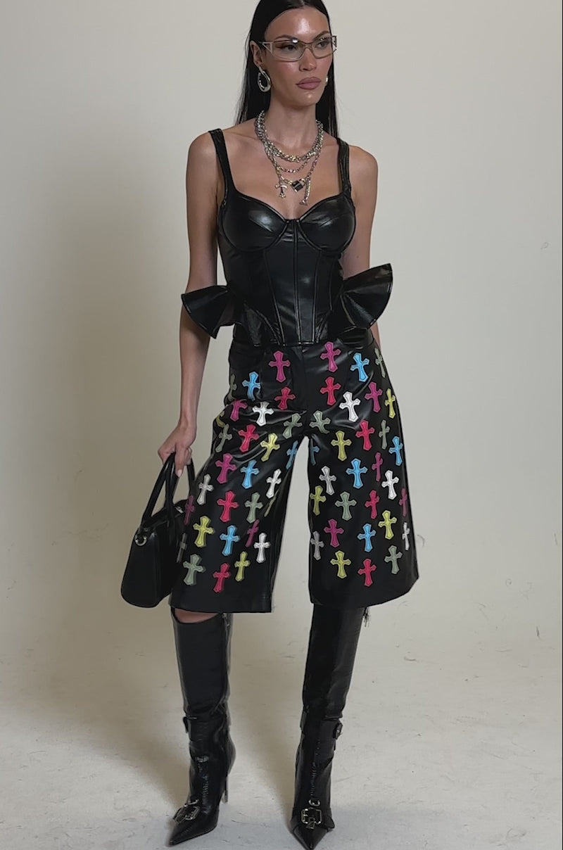 A video of a woman wearing the SEXY AS CAN BE FAUX LEATHER CORSET, colorful cross-patterned black shorts, knee-high black boots, layered necklaces, glasses, and holding a black handbag stands against a plain background.