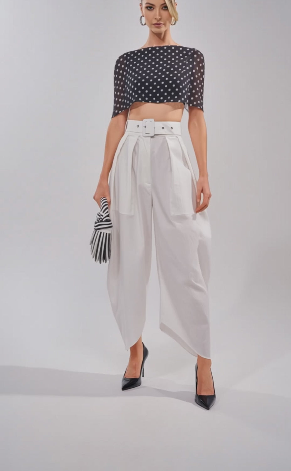 A video of a woman wearing the GIVE ME YOUR LOVE TROUSER—high-rise, white barrel pants with a belt—styled with a sheer black polka-dot cape top, crop top, black heels, striped clutch, and hoop earrings.