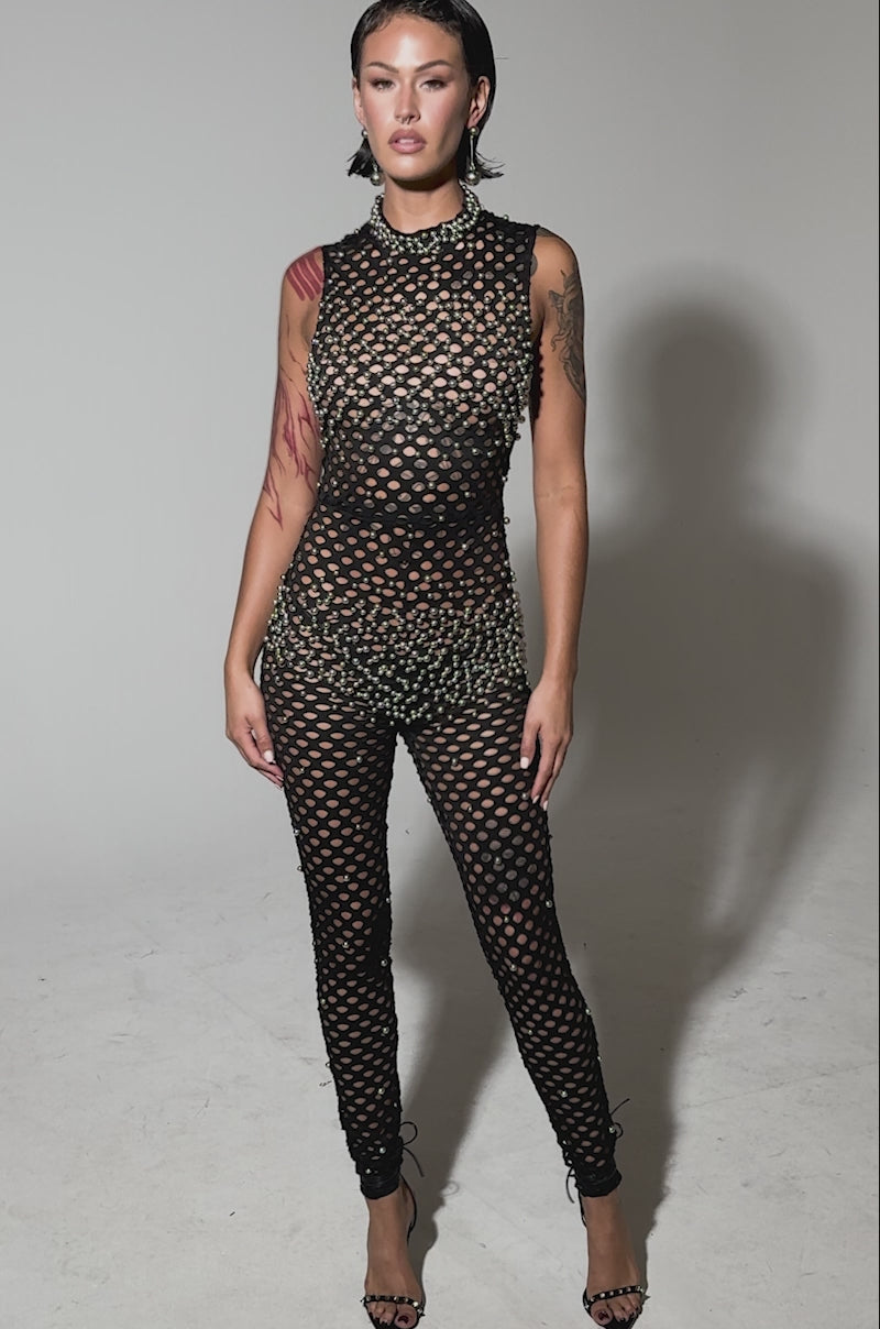 Video of woman modeling the COULD NEVER BE ME BEADED MESH JUMPSUIT, a fitted, sleeveless mesh piece with circular beaded designs, styled with high heels against a plain backdrop.