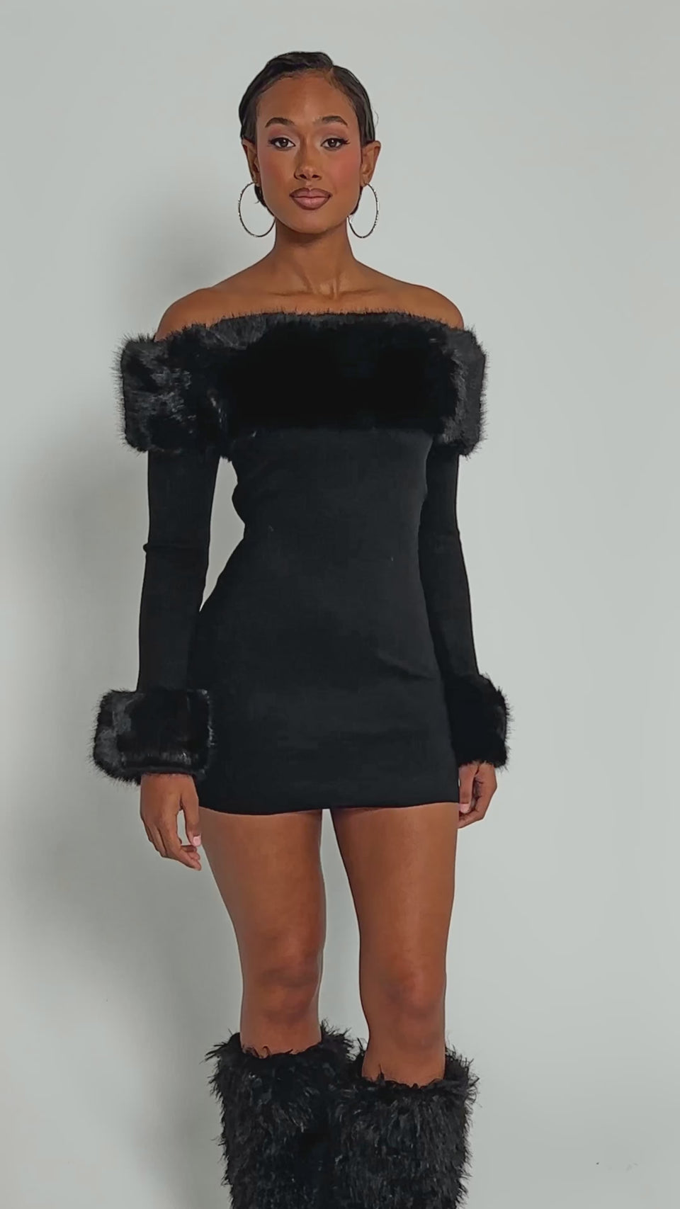 Video of woman modeling the COMFY COZY FUR TRIM MINI DRESS IN BLACK with off-the-shoulder style and faux fur accents, a woman poses with clasped hands, her large hoop earrings shining against a plain background.
