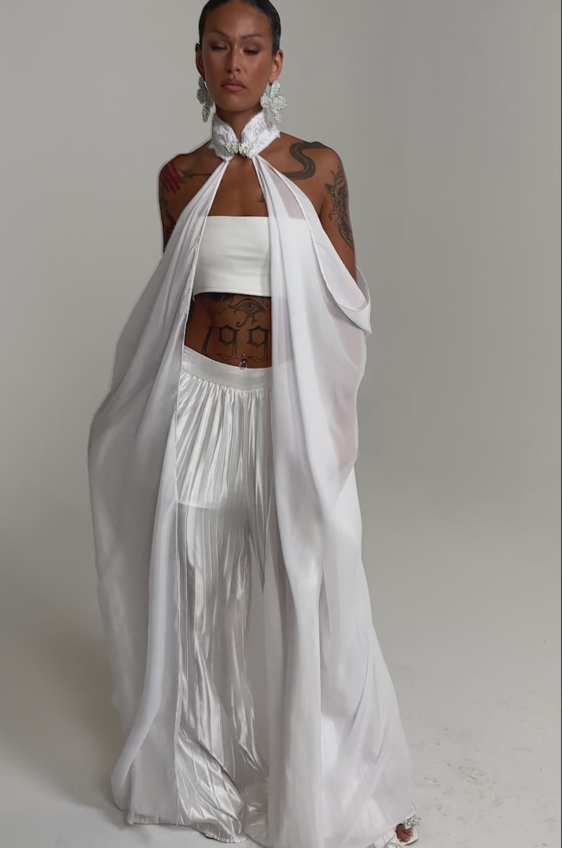 A video of a person wearing the MAKE A STATEMENT CAPE over a white bandeau top and wide satin trousers, styled with tattoos, and statement earrings.