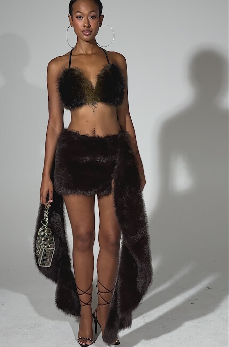A video of a woman posing confidently in the ITS GIVING FUR MINI SKIRT, paired with a black faux fur bikini top, silver chain mini purse, hoop earrings, strappy black heels, and a faux fur sash—an eye-catching look for standout fall fashion.