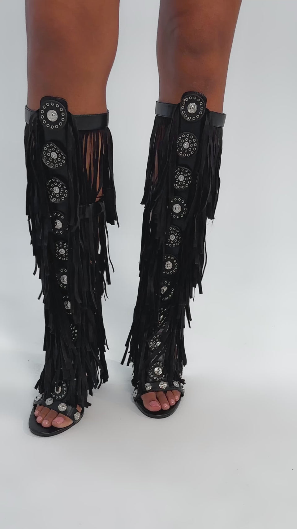Video of woman modeling the AZALEA WANG SAMEYANA BLACK FRINGE AND METAL STILETTO GLADIATOR SANDAL, featuring knee-high black fringe and circular silver accents, standing against a plain light background.
