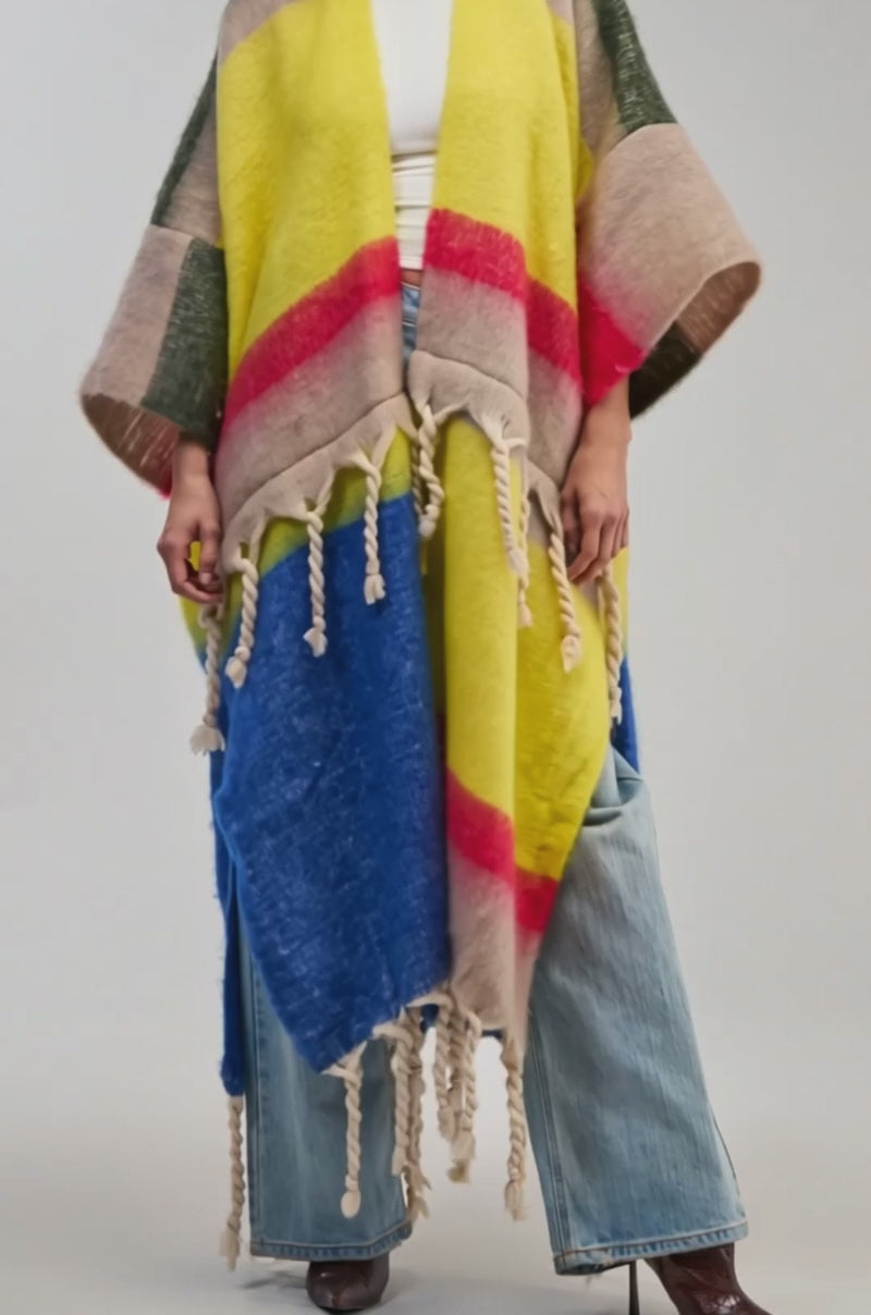 Video of a woman modeling the SUPER COZY OVERSIZED CARDIGAN IN YELLOW MULTI—a vibrant, colorful piece featuring yellow, red, blue, beige, and green sections reminiscent of an oversized fringe sweater.
