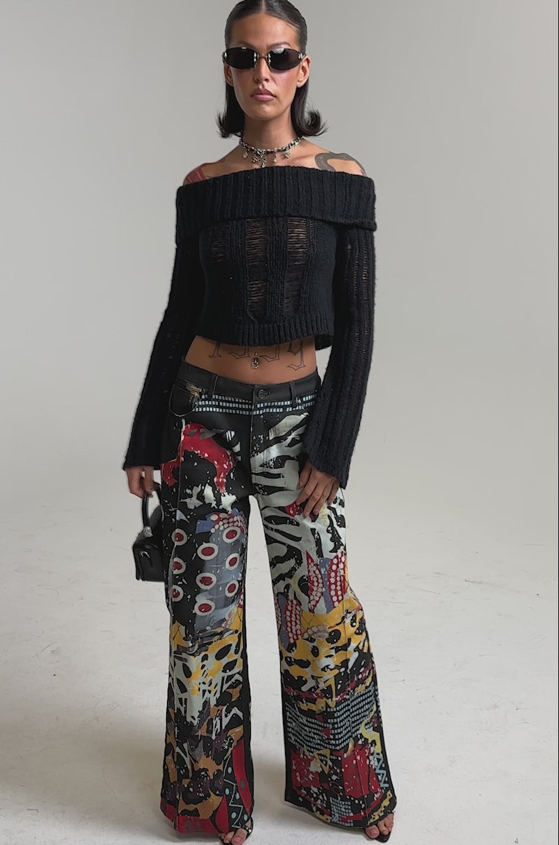 A video of a woman wearing sunglasses, a black off-the-shoulder cropped sweater, and SOMETHING DIFFERENT PRINTED BLACK WASH JEANS with an abstract patchwork pattern, holding a small black handbag.