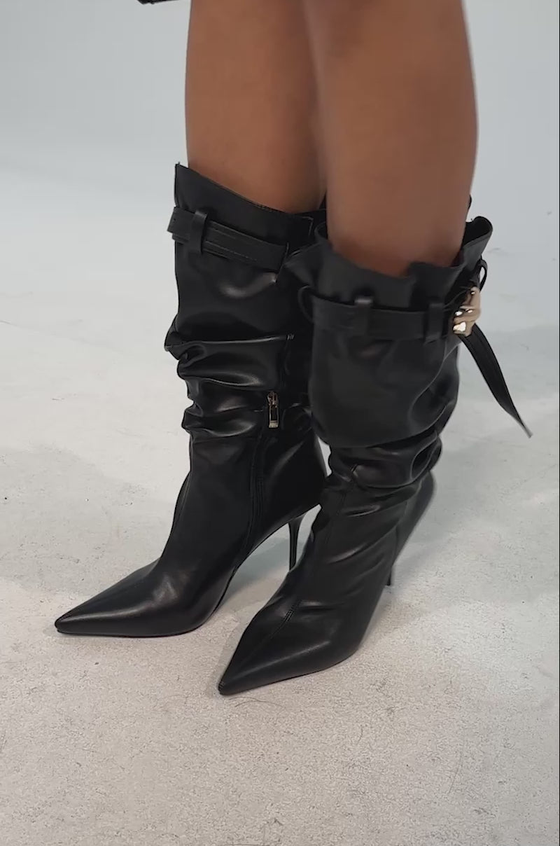 Video of woman modeling the AZALEA WANG AZELINE BLACK PU SLOUCHY STILETTO BOOTS, featuring pointed toes, knee-high slouchy style, gold buckles, and decorative straps, shown from the side.