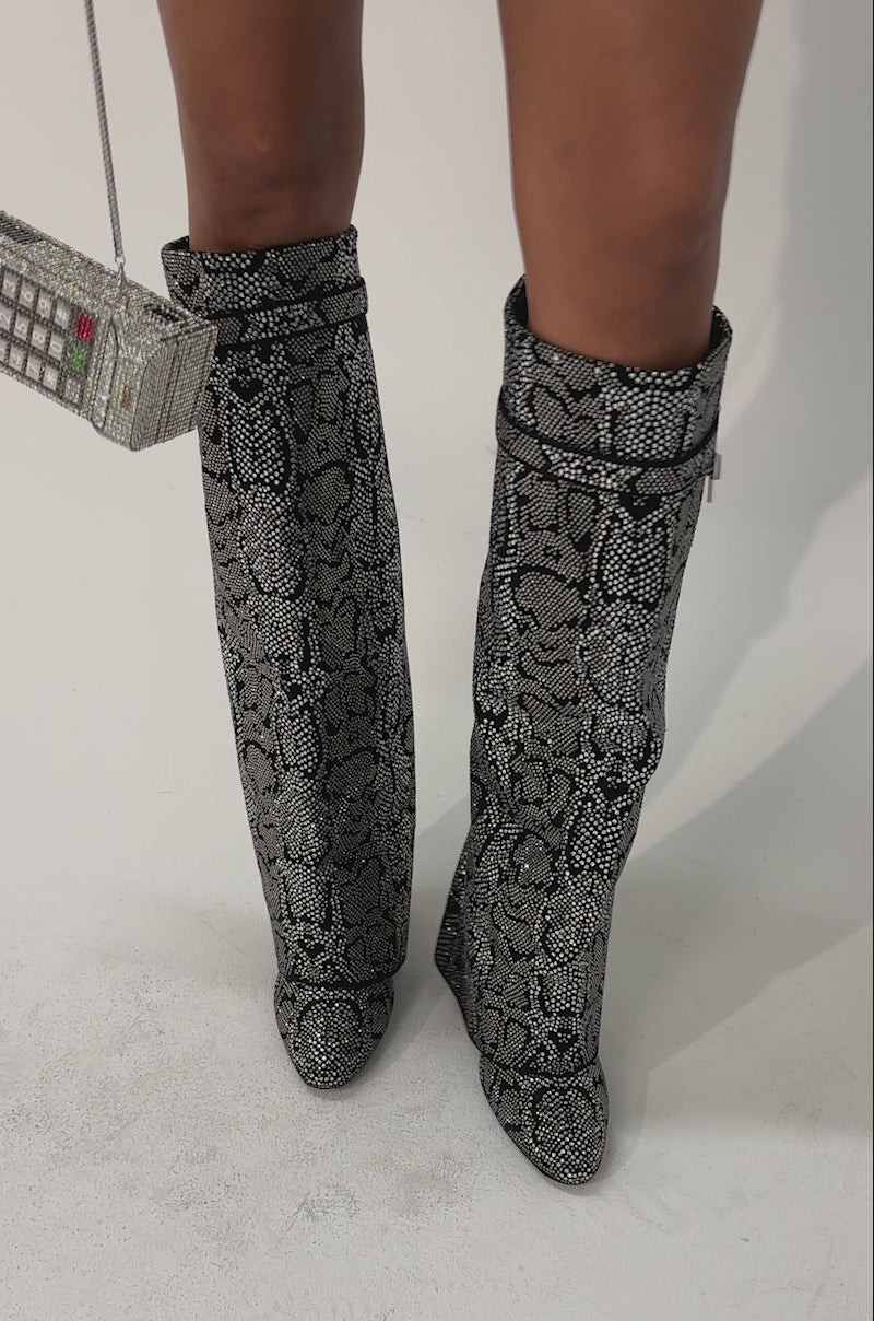 Video of woman modeling the AZALEA WANG CAPRICORN RHINESTONE SNAKE PATTERN WEDGE FOLD OVER BOOT—knee-high, black and gray snakeskin with a loose, slouchy fit, pointed toes, and wedge heels—while standing on a light surface.