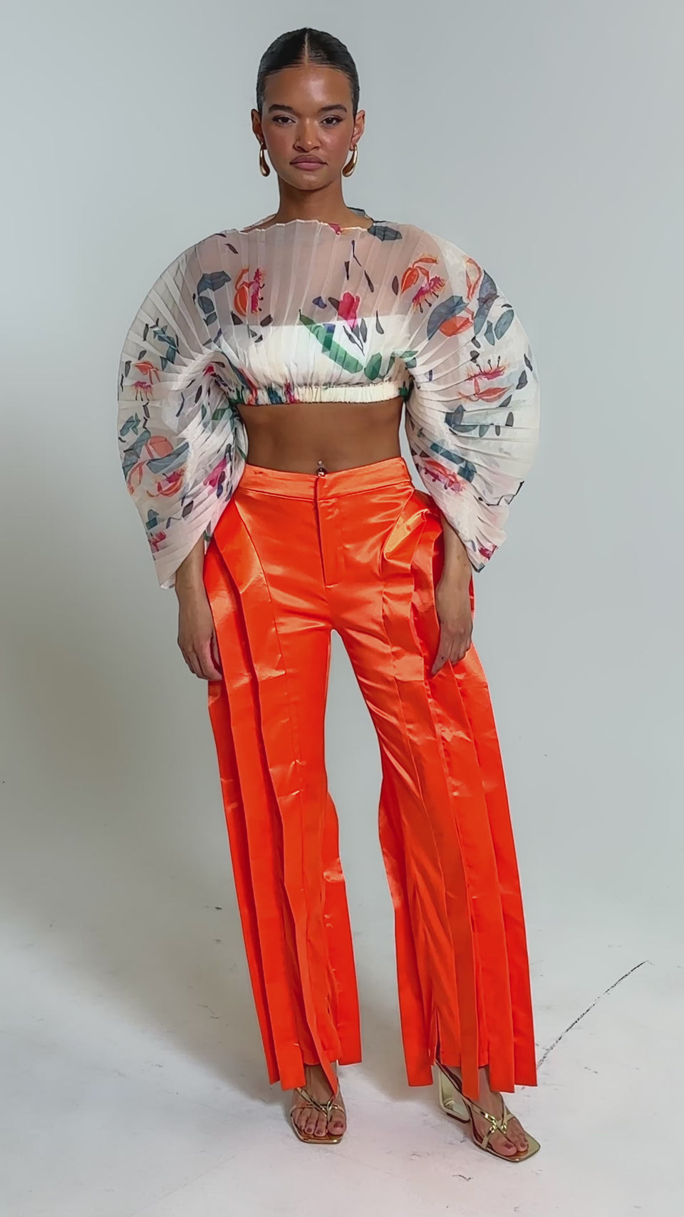 Video of woman modeling the CANDY RAIN PLEATED BLOUSE with colorful abstract patterns and sculptural sleeves, paired with bright orange pleated pants—a striking choice for chic occasions.