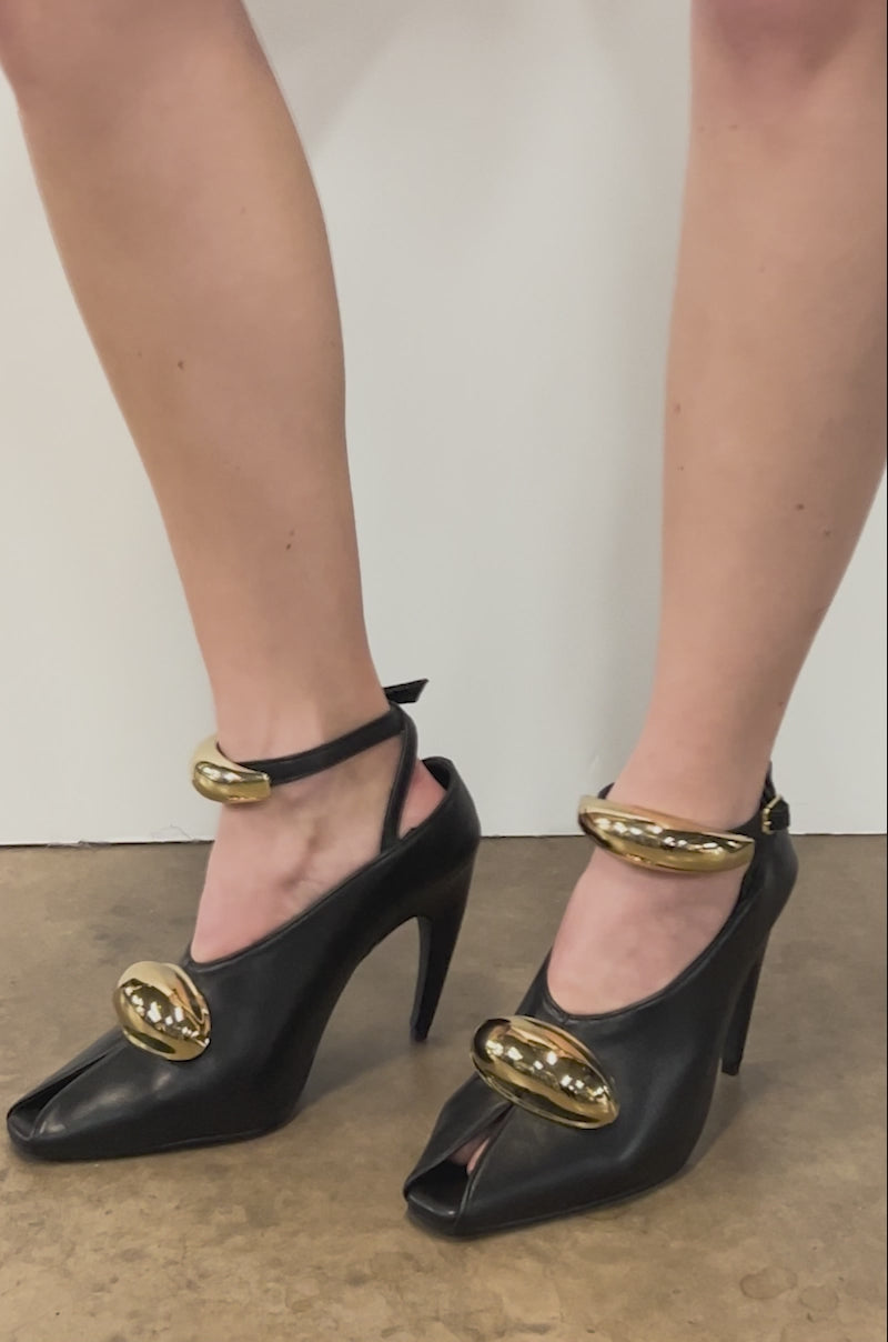 A close-up video of feet in AZALEA WANG HIMAH BLACK OPEN TOE PUMP heels featuring a split-toe design and two bold gold oval accents at the ankle and forefoot, standing on textured concrete.