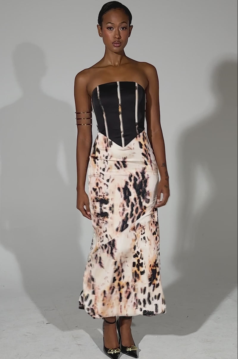 A video of a woman modeling THE REAL DEAL PRINTED MAXI DRESS, featuring a black corset-style satin top and flowing animal-print maxi skirt. She pairs it with metallic heels, a gold arm cuff, slicked-back hair, and standing before a plain light background.