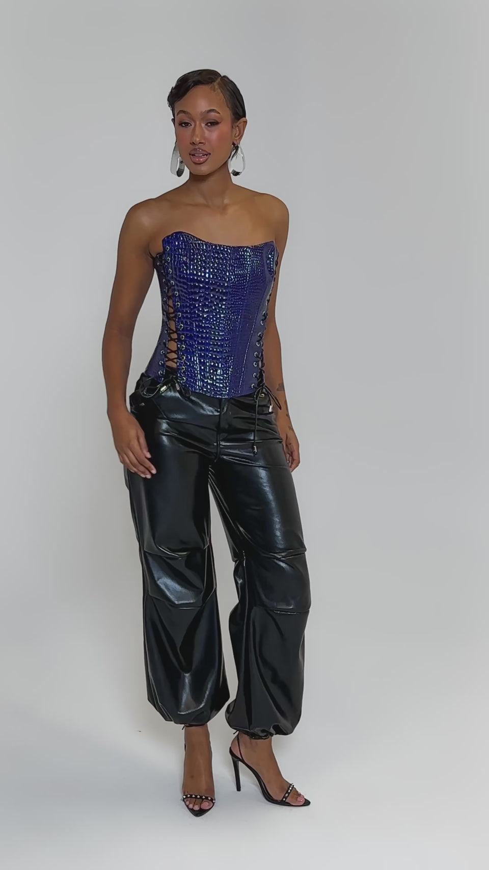 Video of woman modeling the HIT THE SWITCH EMBOSSED CORSET, a blue strapless crocodile print top with lace-up sides, paired with black faux patent leather pants and large geometric earrings, posing against a plain light background.