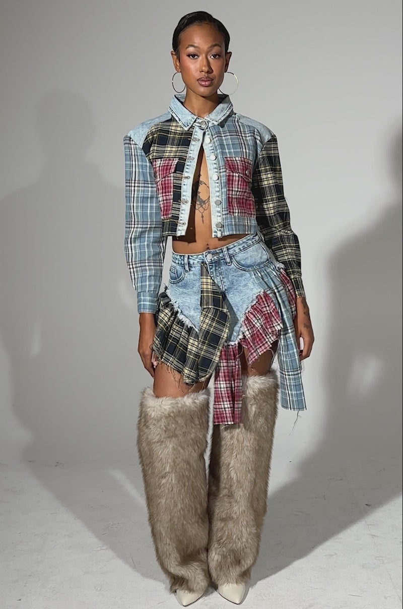 A video of a person posing confidently in the AUTUMN VIBES PLAID MINI SKIRT, paired with a patchwork jacket, large hoop earrings, and tall furry beige boots. The bold look pops against a plain light background.