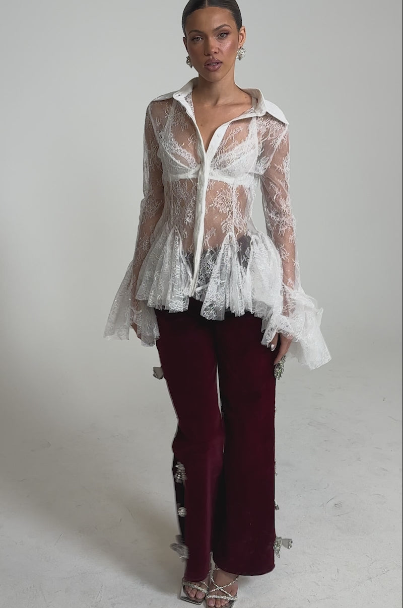 A video of a woman wearing the LADY LOVER LACE BUTTON DOWN, a sheer lace blouse with flared cuffs, styled with burgundy velvet pants featuring decorative accents and strappy high-heeled sandals.