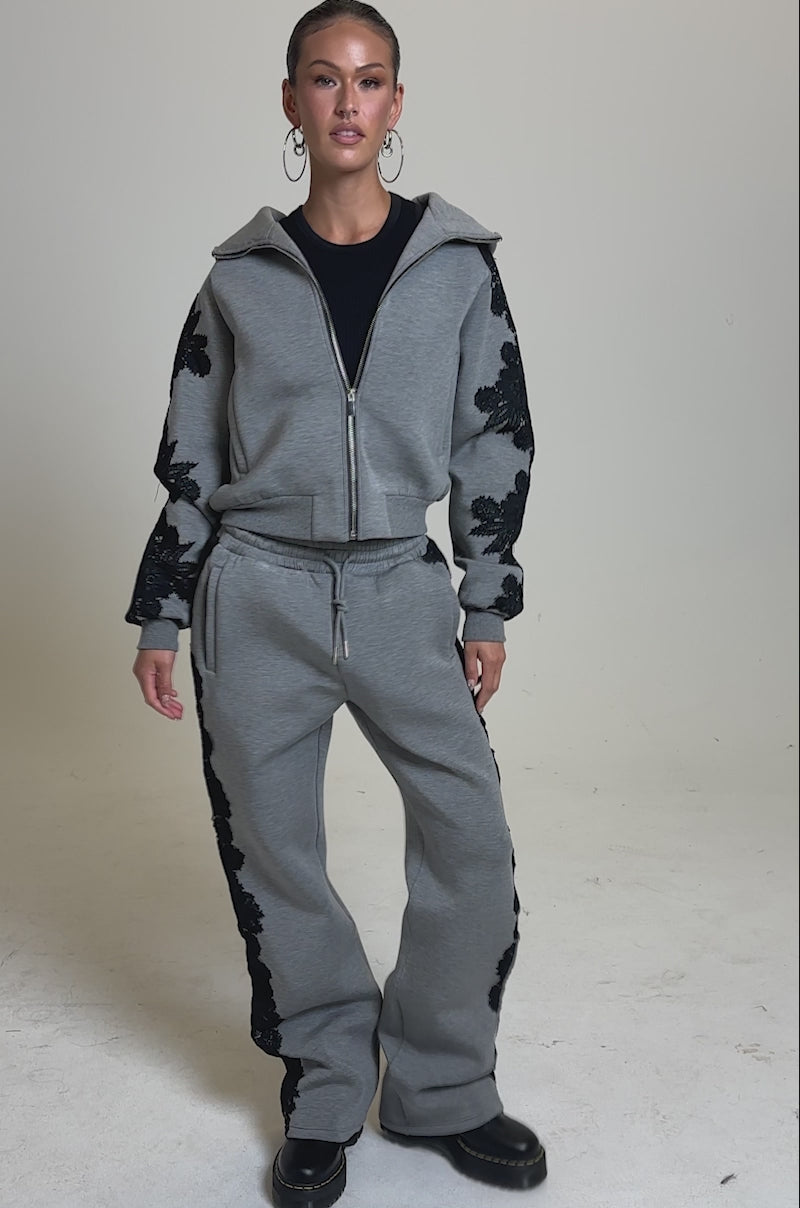 A video of a woman wearing the RAQUEL STRAIGHT JOGGER PANT, a gray high-rise stretchy pant with black floral lace appliques, paired with a matching zip-up jacket, black top, hoop earrings, and black platform shoes against a plain background.