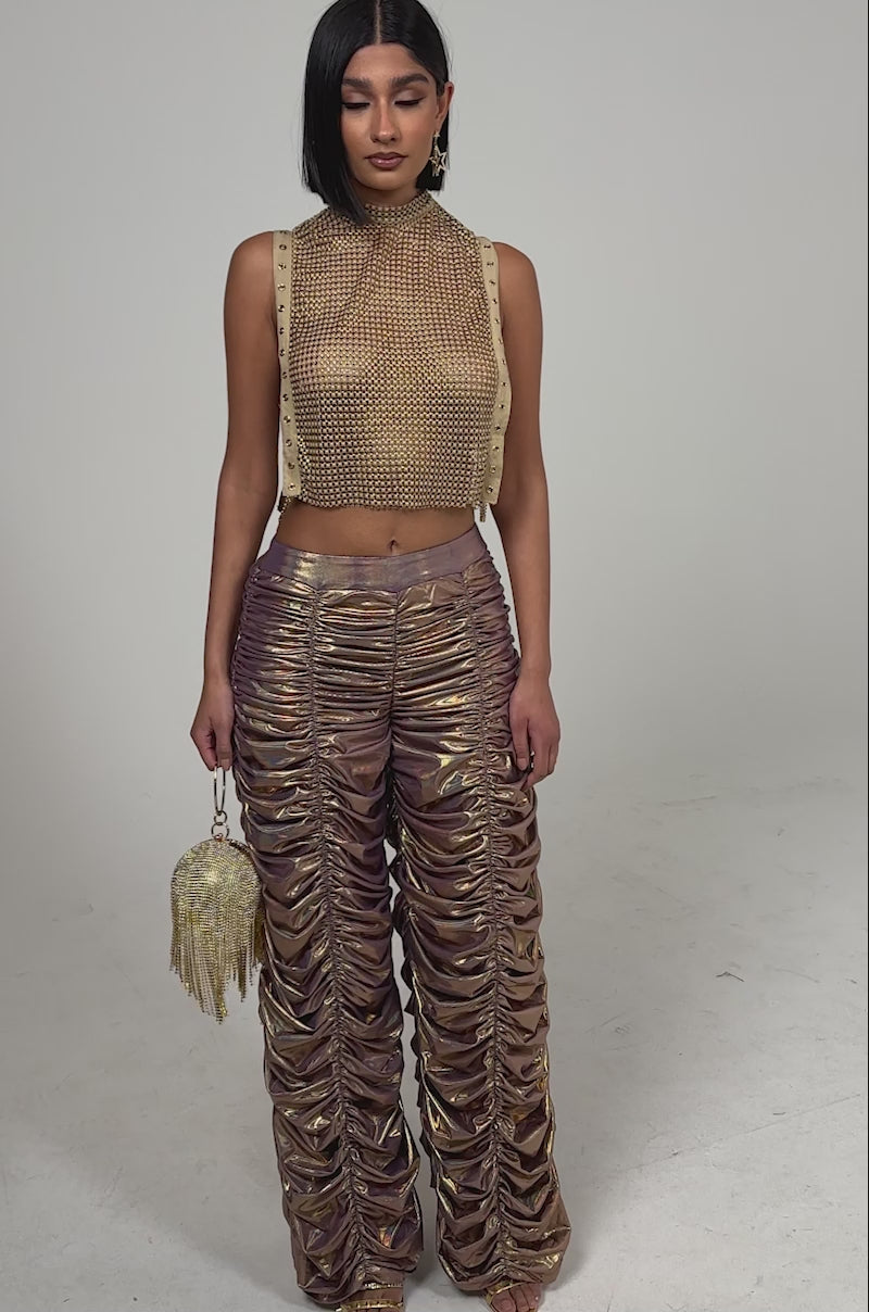 A video of a woman in the TEARS IN THE CLUB OPEN SIDES MOCK NECK RHINESTONE MESH BLOUSE paired with shiny bronze ruched pants, a round gold tassel purse, gold earrings, and heels against a plain light background.