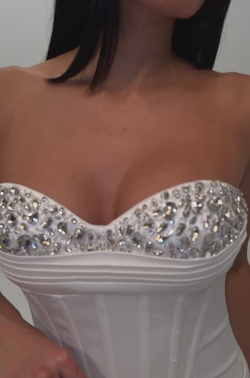 A video of a woman wearing the CRUSH HOUR EMBELLISHED SATIN MINI IN WHITE, featuring a rhinestone-embellished design and jeweled, structured bodice. 