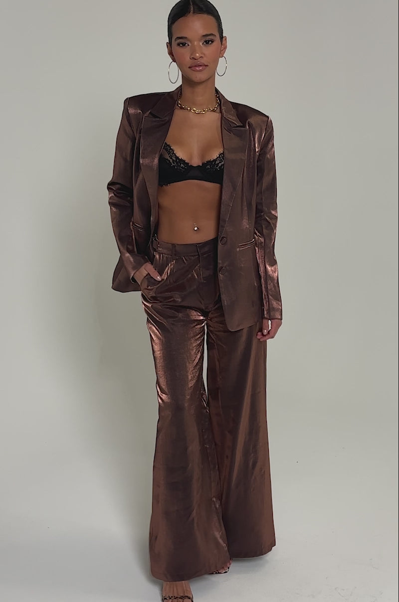 Video of woman modeling the STAY CLOSE IRIDESCENT BLAZER over a black lace bralette, she completes her look with slicked-back hair, large hoops, and a gold chain necklace as she poses against a plain light background.
