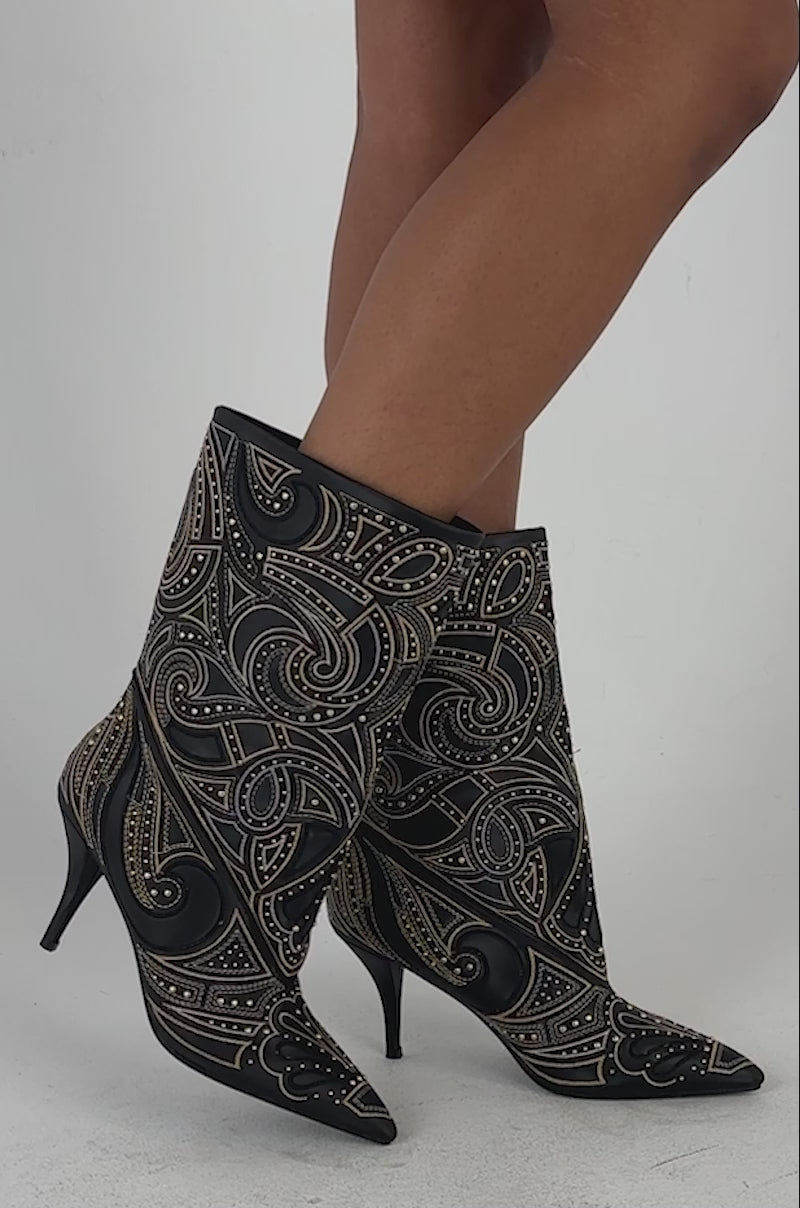 Video of a woman modeling the AKIRA GOLD ZORINA BLACK MULTI METALLIC PATTERNED BOOTIE sits on a clear chair; only their legs and the striking multi metallic patterned boots are visible.
