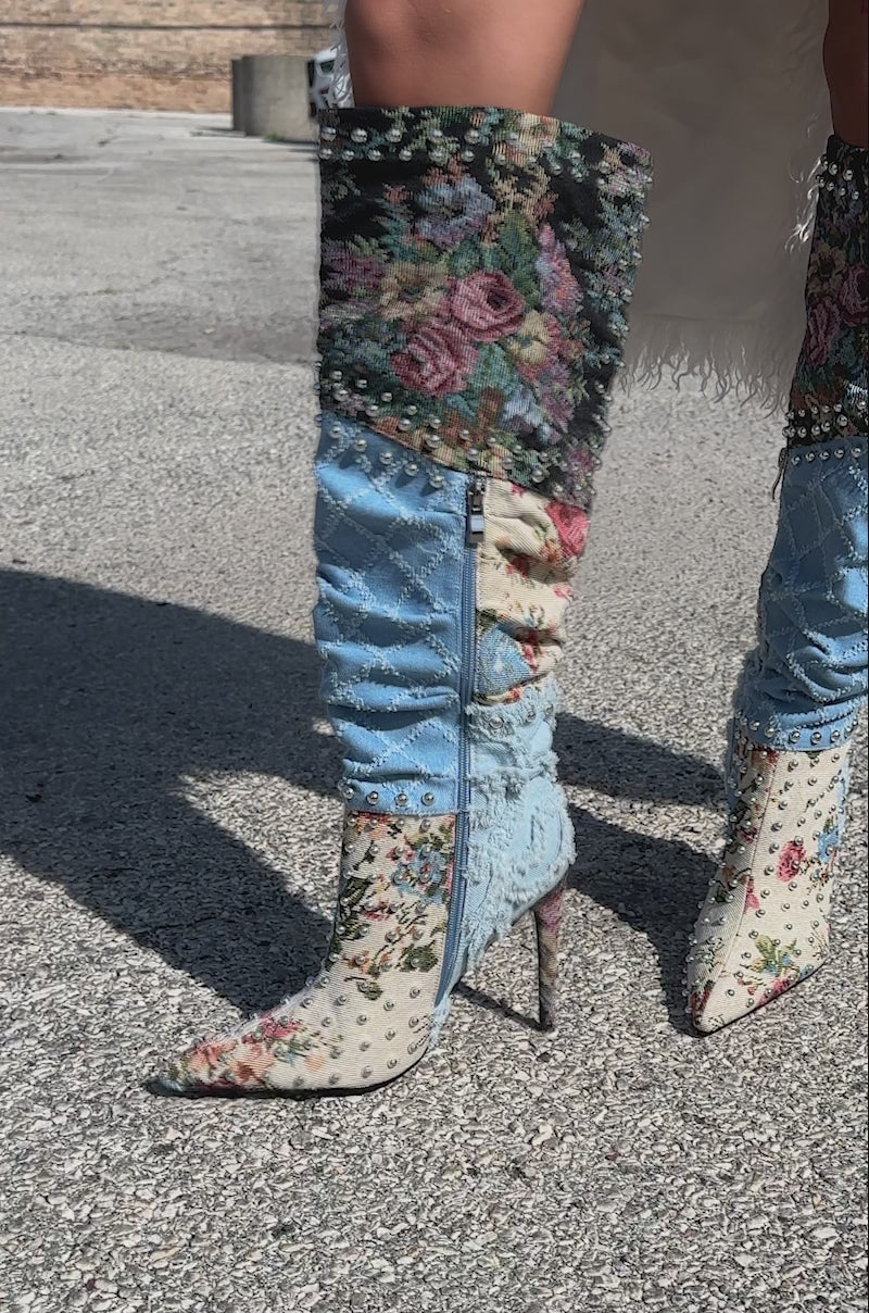 A video of a person wearing the AZALEA WANG AILA DENIM MULTI STUDDED POINTED TOE STILETTO BOOT, styled with a cream ruffled skirt and showing a thigh tattoo, stands on a paved surface.