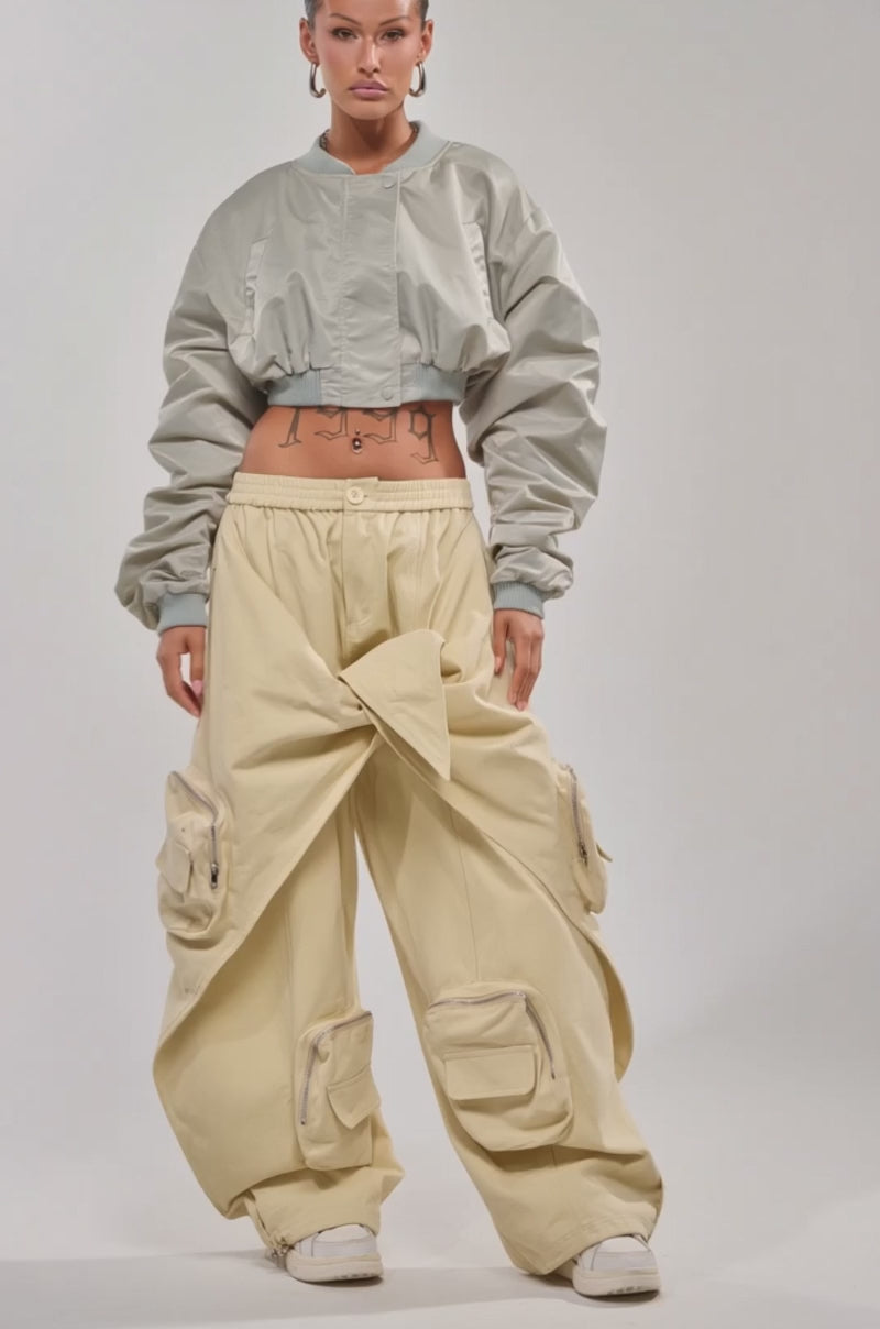 Video of woman modeling the TAKE IT EASY CROPPED BOMBER IN SAGE, beige wide-leg pants with a knotted front, large hoop earrings, and showcasing visible tattoos on her midriff while posing confidently against a plain background.