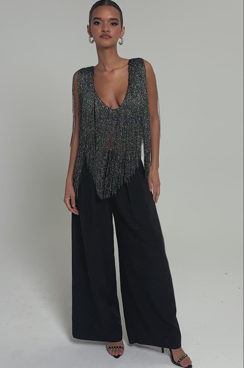 Video of woman modeling the DEVVON FRINGE TOP featuring metallic threads and a deep neckline, paired with black pants, standing against a plain background.