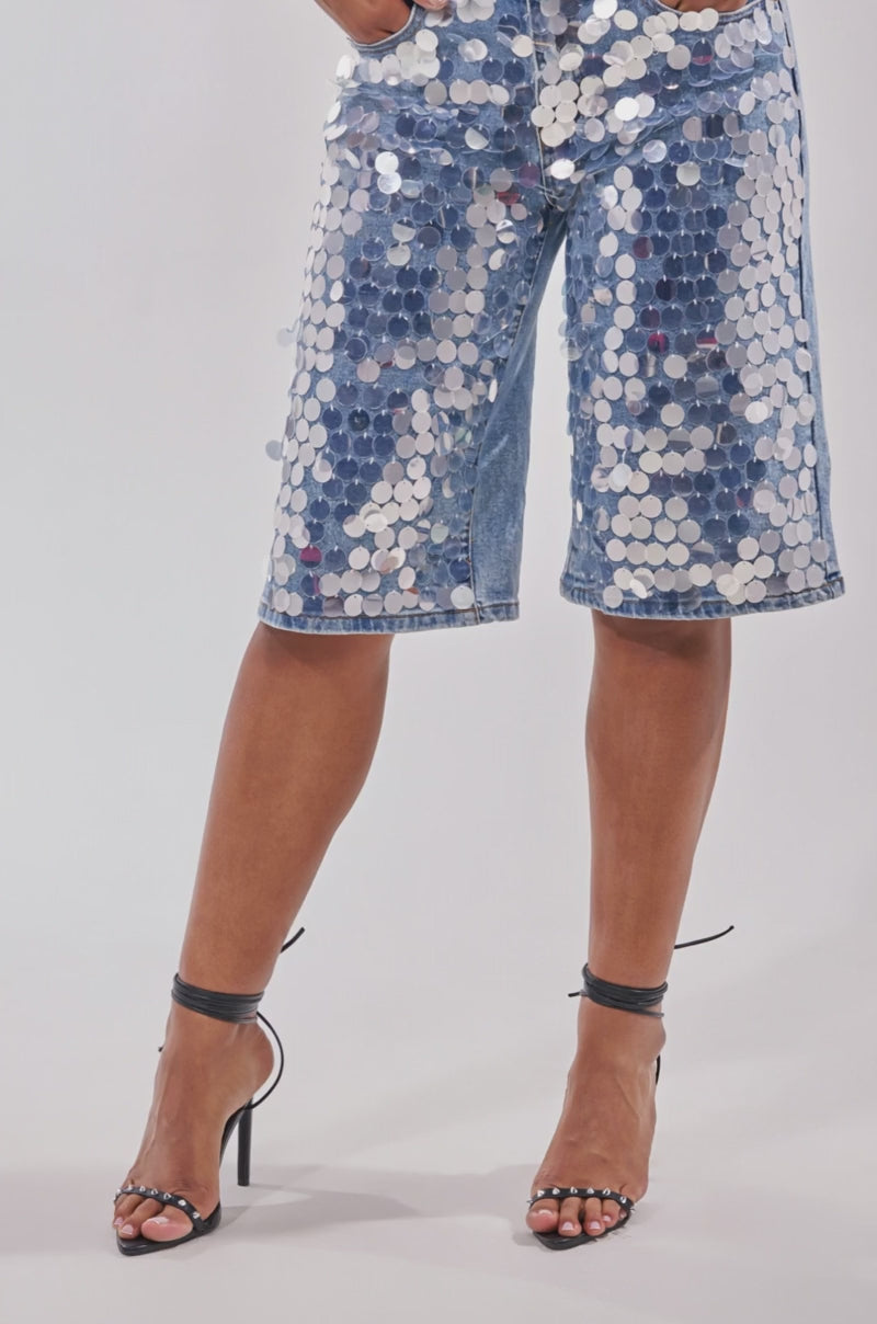 A video of a model earing the SPARKLE AND SHINE BERMUDA SHORT, adorned with large silver sequins, and black strappy heels.