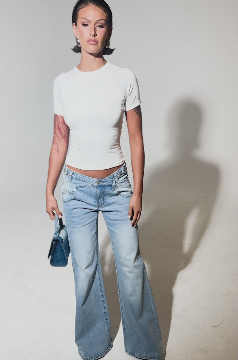 A video of a woman standing against a plain background in a fitted white short-sleeve top, gray shoes, and the WON'T LOSE MYSELF WIDE-LEG BLUE DENIM jeans. She holds a small blue handbag and has tattoos on her arms.