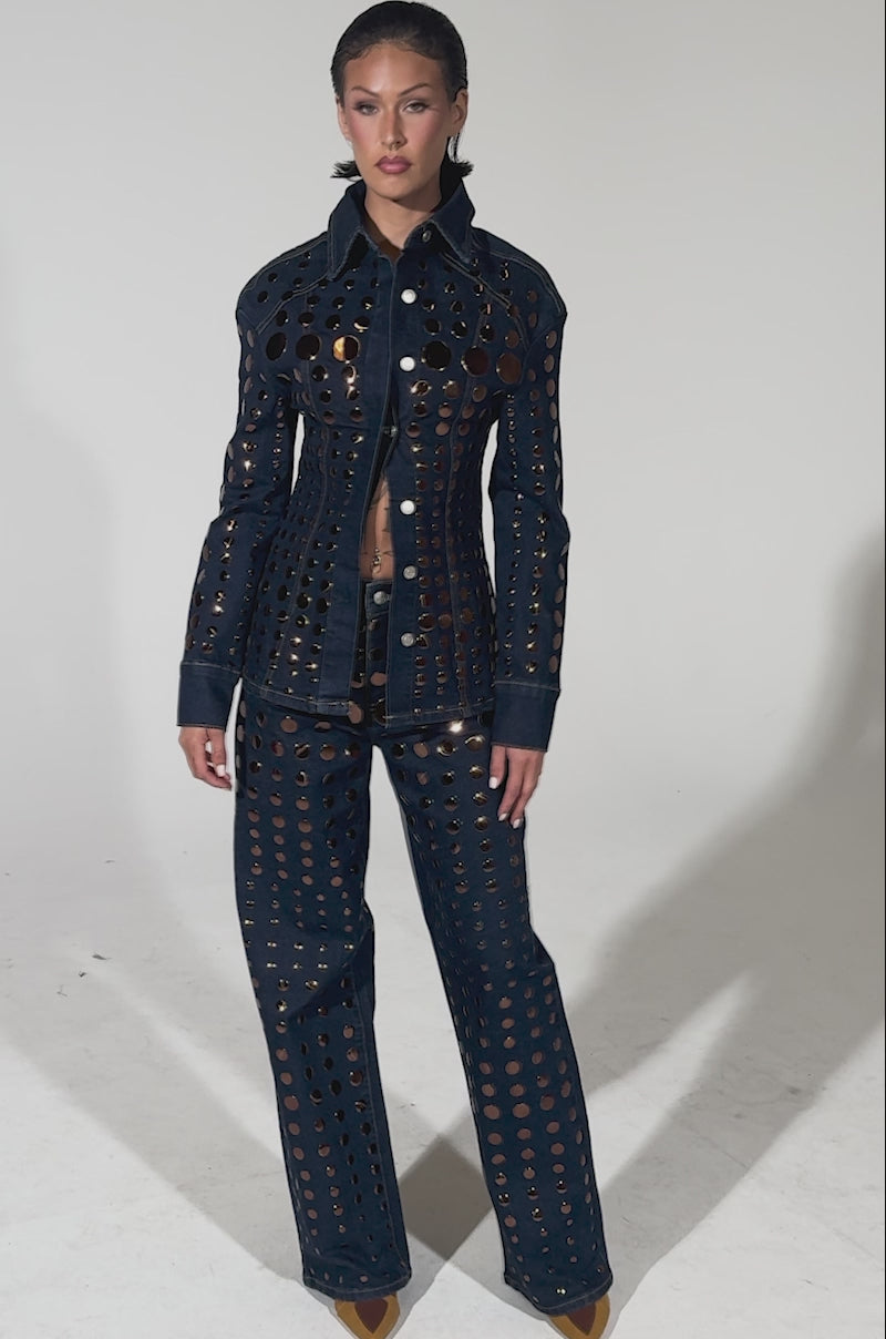 A video of a person wearing the DISCO BABE RELAXED DENIM set—dark blue, high-waisted denim jacket and wide-leg jeans with copper eyelet details—paired with tan shoes, standing before a plain light gray background.