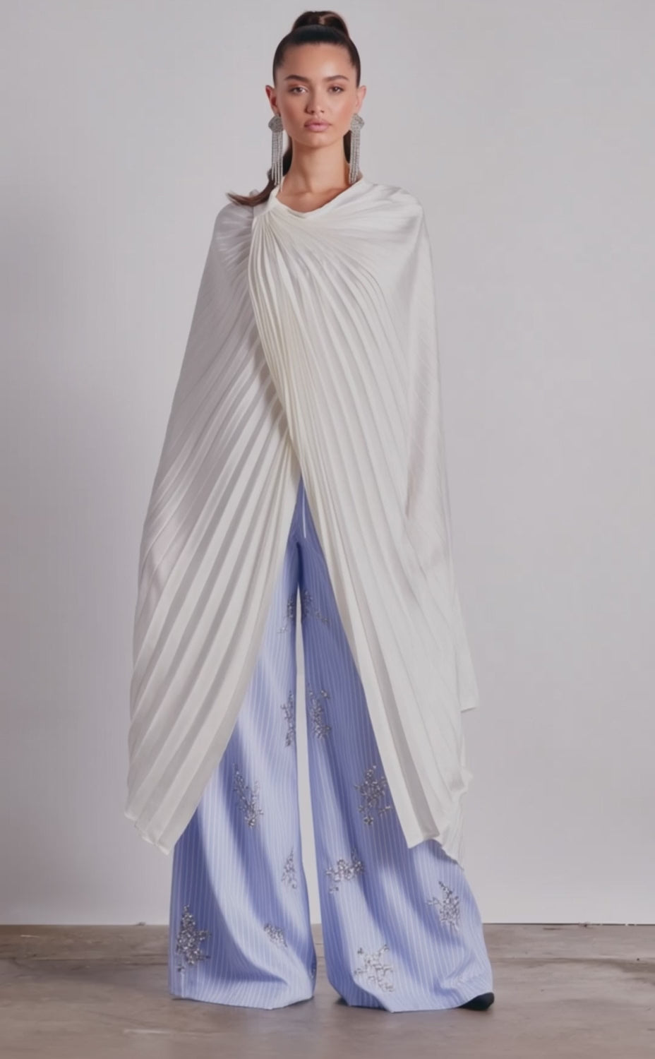 A video of a person wearing the FARAH DRAPED PLEATED CAPE TOP IN WHITE over wide-legged, light blue pants with silver embroidery. 