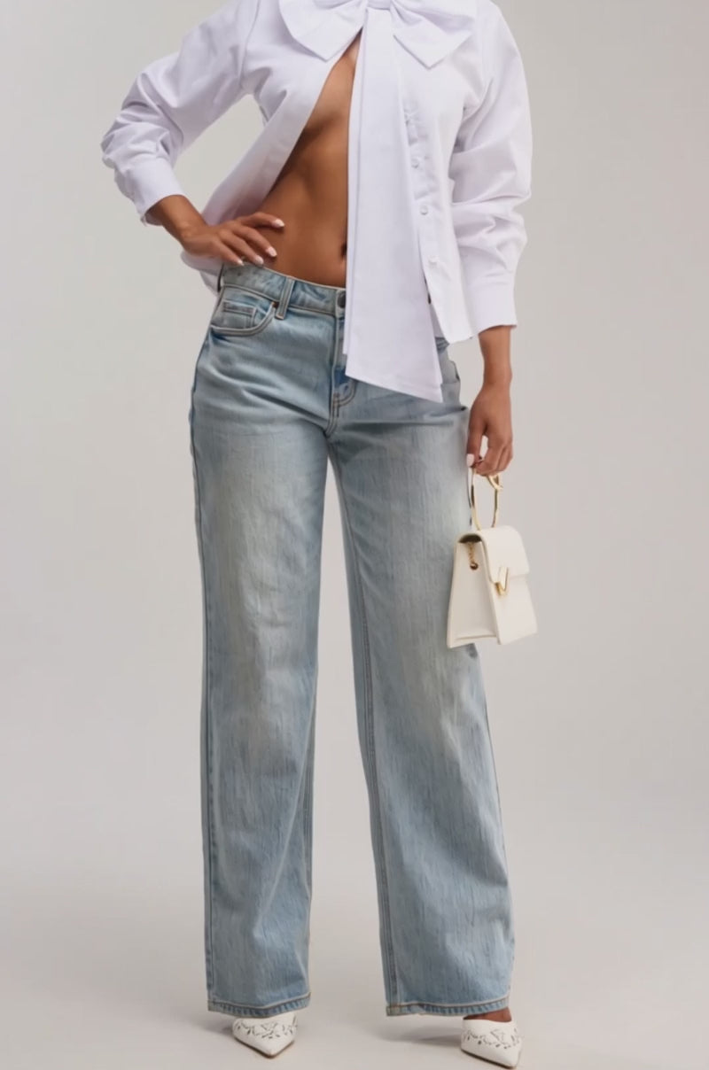 Video of woman modeling the BUSINESS OR PLEASURE POPLIN BLOUSE, featuring an oversized bow at the collar, paired with light blue jeans, gold heart-shaped earrings, and holding a white handbag with a gold handle.