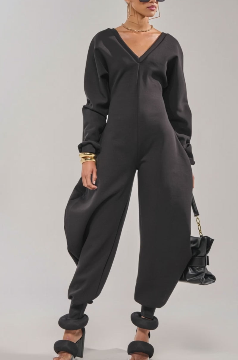 A video of a woman in the COMFY COZY SWEATSHIRT JUMPSUIT IN BLACK, styled with black heeled sandals, sunglasses, gold jewelry, and a black handbag with a gold chain strap against a plain light background.