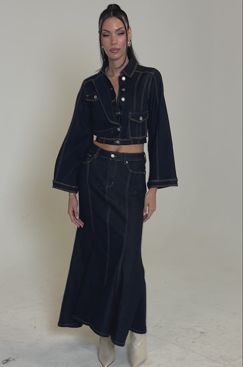 A video of a woman modeling the SHES GONE, DANCE ON DENIM MAXI SKIRT with a dark denim cropped jacket featuring flared sleeves and white buttons, styled with white pointed-toe boots and sleek, brushed-back hair.