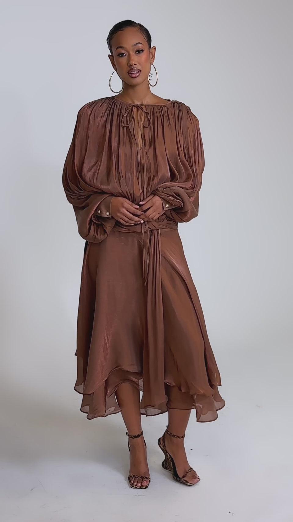 A video of a woman in the GINA MAXI DRESS, a shiny copper dress with layered, flowy fabric and a flattering waist tie. 